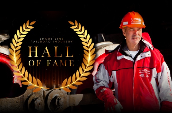 Rick Corman wearing a red R. J. Corman coat and orange hard hat next to the ASLRRA Short Line Railroad Industry Hall of Fame seal.