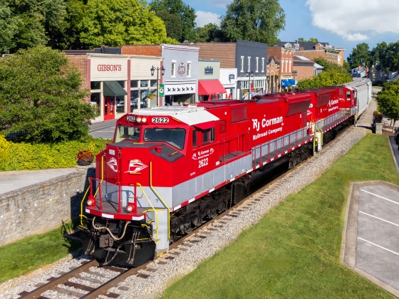 A red R. J. Corman locomotive passing a row of buildings. 