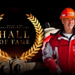 Rick Corman wearing a red R. J. Corman coat and orange hard hat next to the ASLRRA Short Line Railroad Industry Hall of Fame seal.