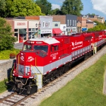 A red R. J. Corman locomotive passing a row of buildings. 